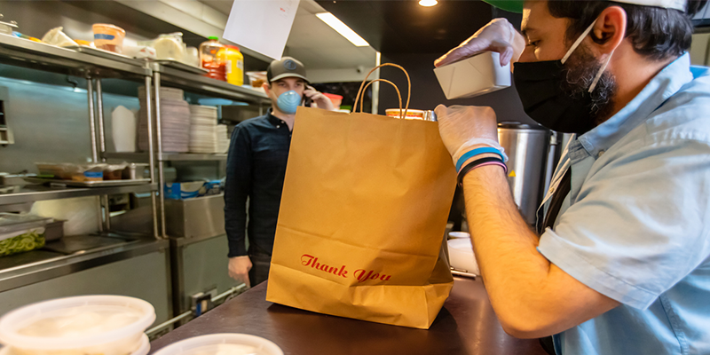 A restaurant worker in a mask prepares a takeout order while another employee stands nearby, surrounded by kitchen items.